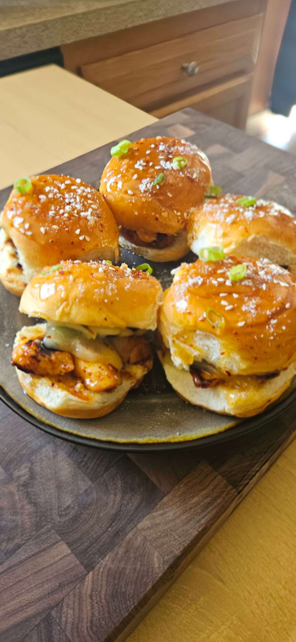 A rustic grey ceramic plate filled with five glossy pull-apart sliders. Each slider is baked on a soft Hawaiian-style bun brushed with garlic-parmesan honey butter, lightly sprinkled with grated parmesan. The chicken is marinated with buttermilk and a special spicy chicken marinate. Pieces of them are sweet-spicy glaze, layered with melted pepper jack cheese and sautéed jalapeños.