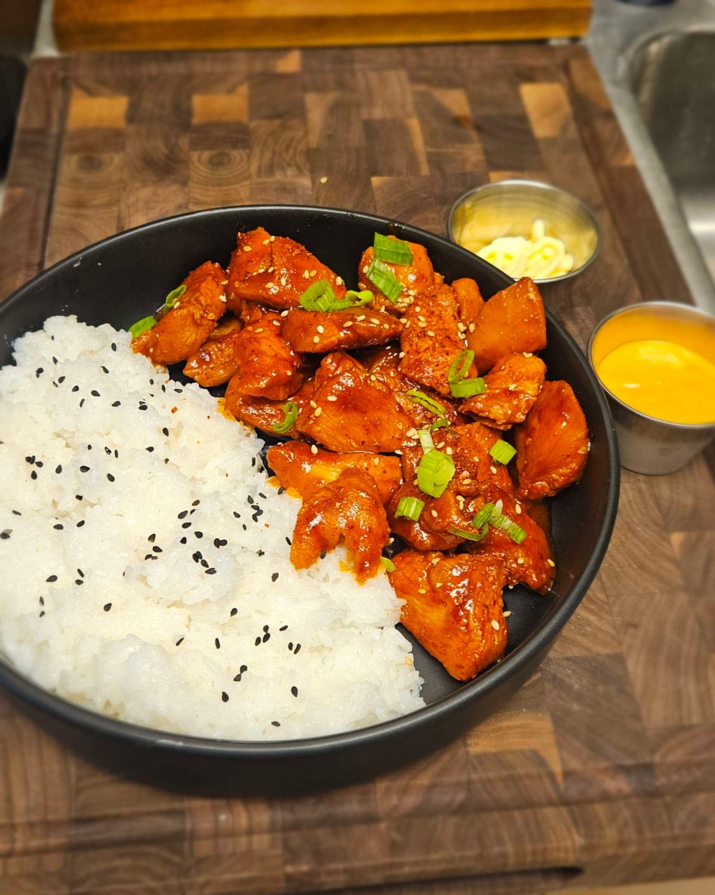 A black bowl filled with spicy Korean chicken, topped with sesame seeds and green onions, served alongside sticky white rice garnished with black sesame seeds. Two small dipping sauces of kewpie mayo and yum yum sauce sit behind the bowl on a wooden surface.