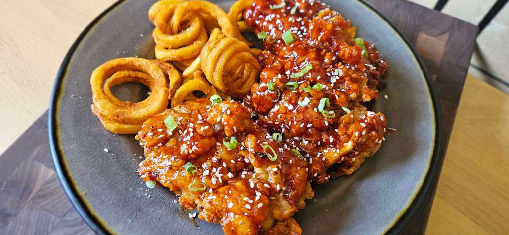 A close-up of a plate featuring crispy buttermilk Korean BBQ chicken glazed in a glossy, deep red sauce. The chicken pieces are generously coated, sprinkled with white sesame seeds, and garnished with finely chopped green onions for a fresh pop of color. Beside the chicken, there’s a portion of golden-brown curly fries, perfectly seasoned and fried to a crisp, their spiral shapes adding a playful contrast to the rich, savory chicken. The food is served on a dark, textured plate with a subtle golden rim, set on a wooden table that enhances the warm, appetizing tones of the meal.