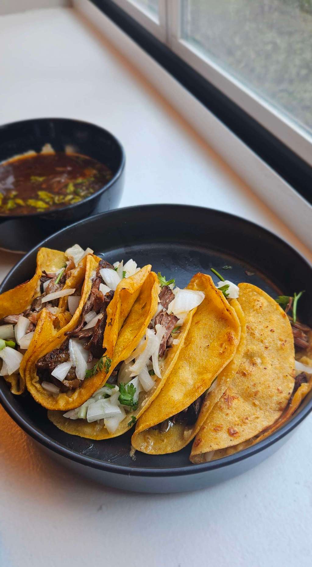 Golden, crispy birria tacos stuffed with tender, slow-braised beef, diced onions, and fresh cilantro, served with a steaming bowl of rich dipping consomé.