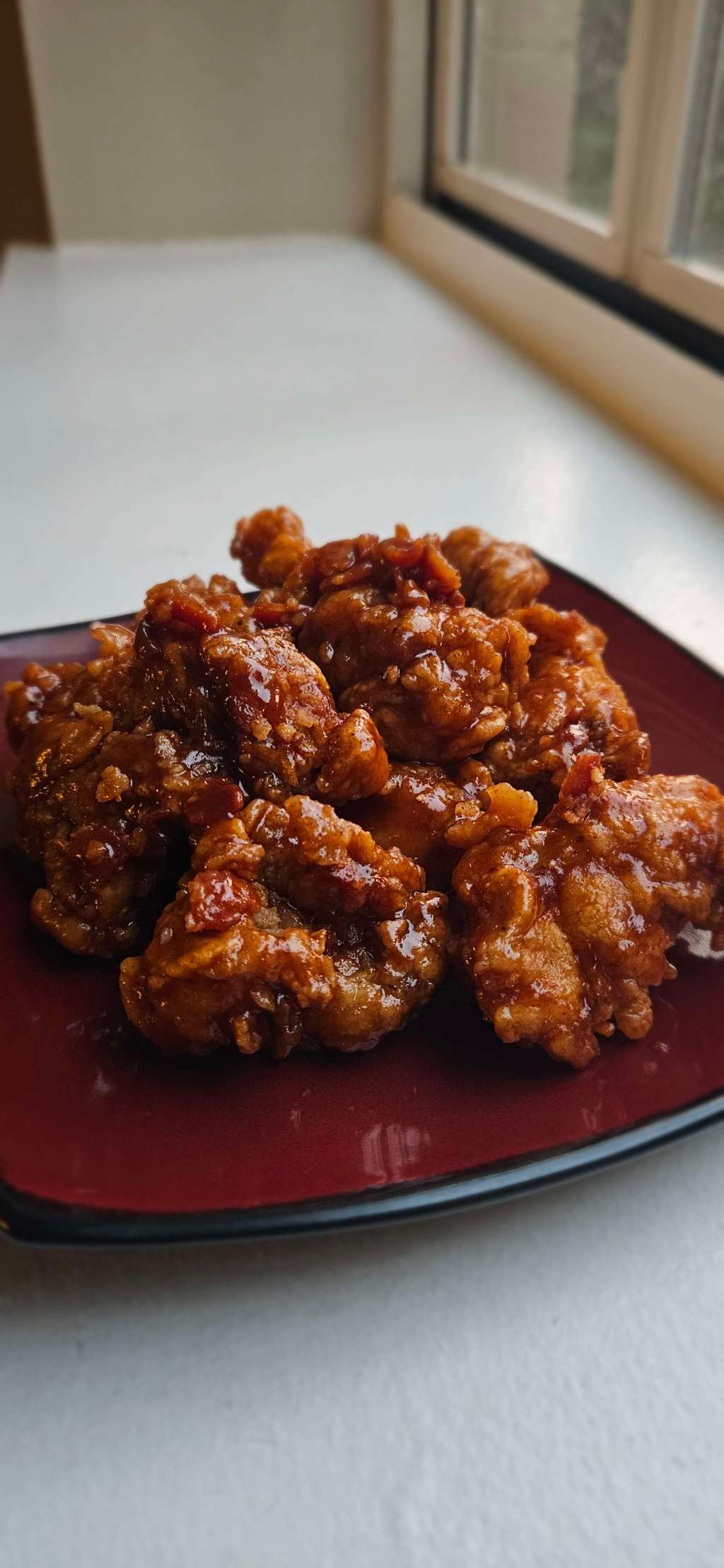 A close-up of crispy fried chicken wings coated in a glossy maple barbecue glaze, piled on a dark red plate by a sunlit window