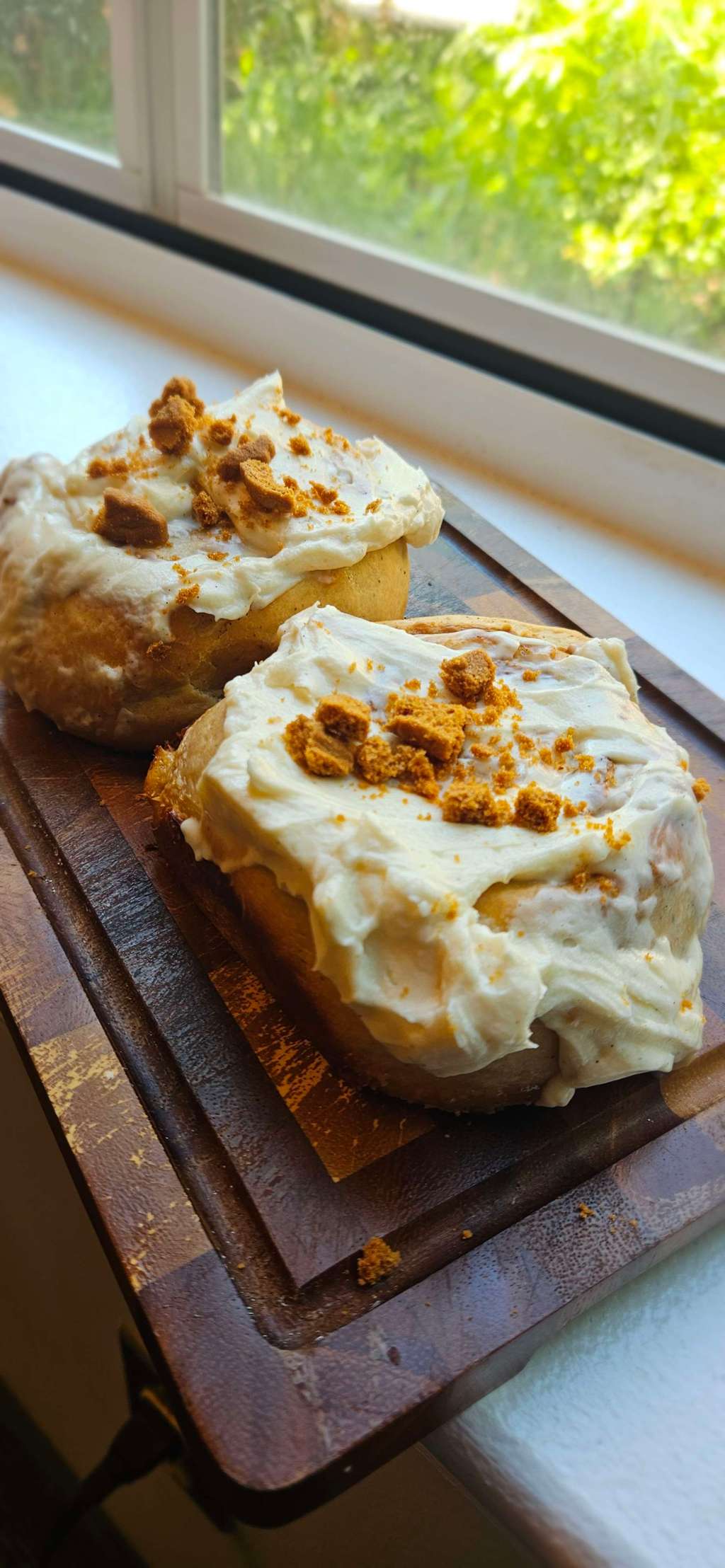 Picture of two jumbo-sized Bischoff cinnamon rolls, layered with cream cheese icing, cookie crumbles, all under a checkerboard wooden cutting board