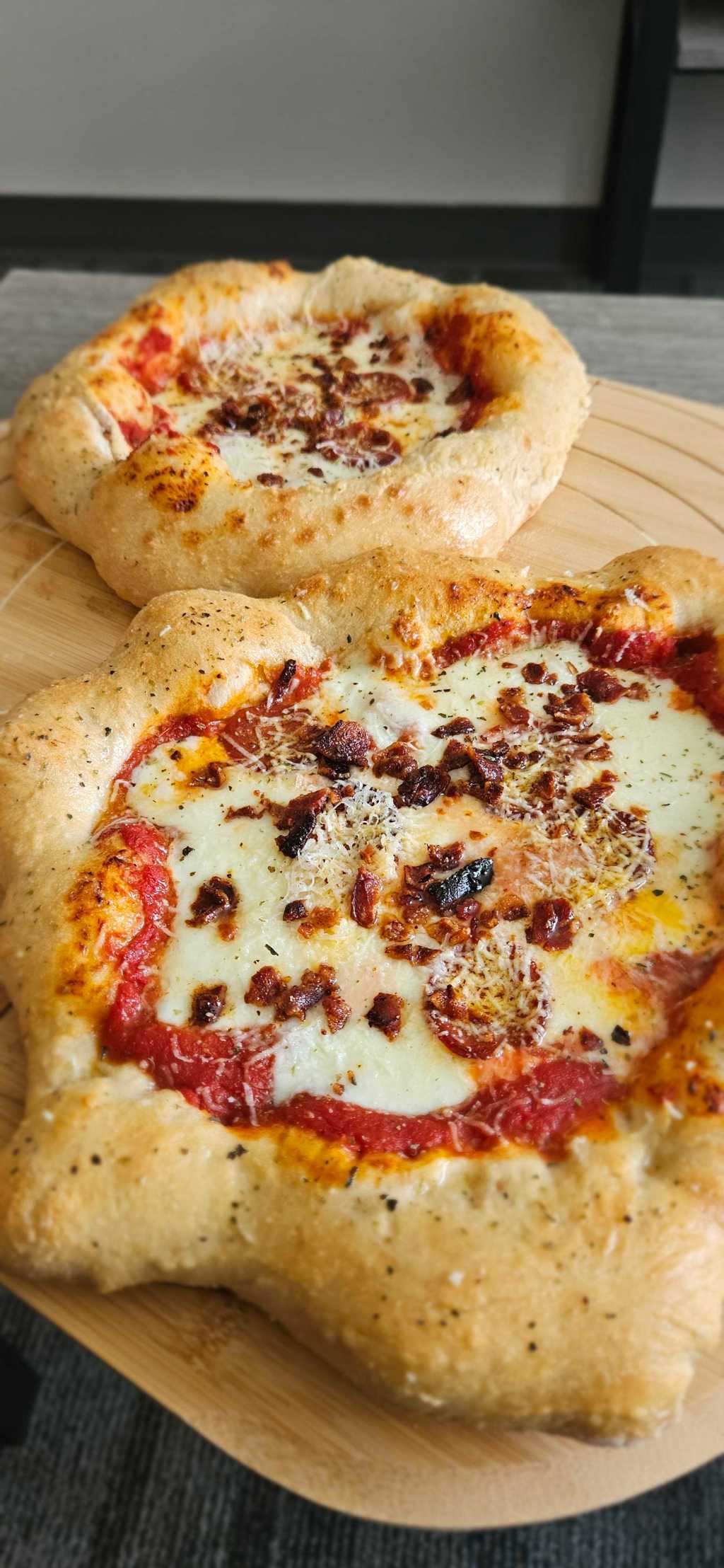 A homemade personal pizza with thick, golden, seasoned crusts sit on a wooden board. They are topped with tomato sauce, melted cheese, bacon pieces, herbs, and grated cheese, with one pizza in the foreground and another slightly blurred in the background.